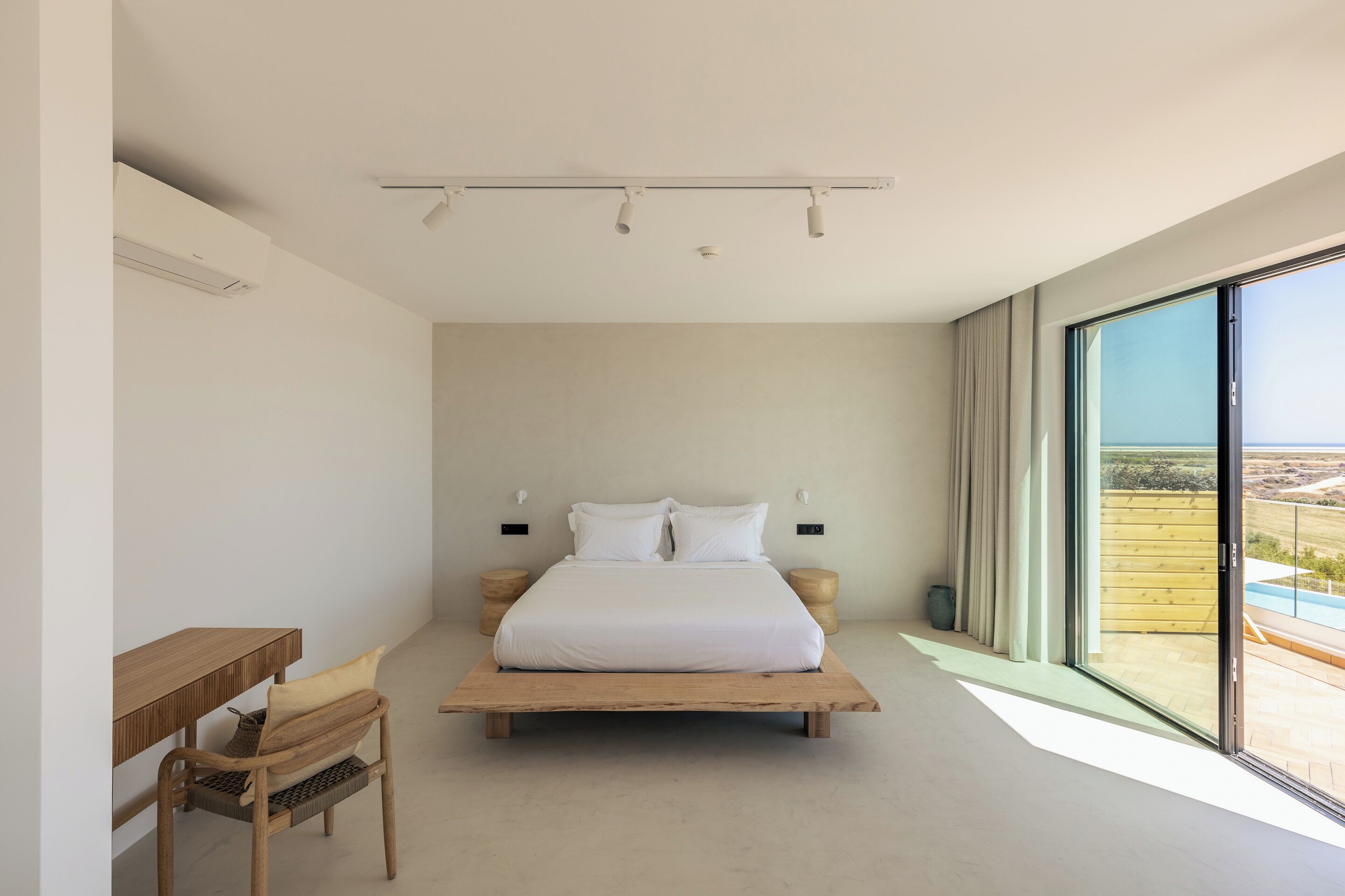 A modern bedroom with a large bed, a wooden desk, and a view of the outdoors.