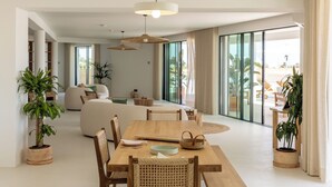 A spacious living area with a wooden dining table, wicker chairs, and a large sofa. There are potted plants and a large window providing natural light. - Villa Loreto (Olhao)