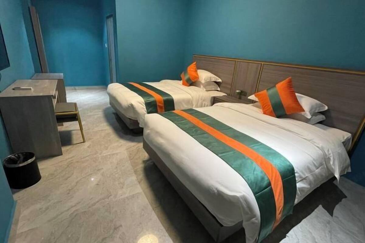 A hotel room with two beds, each with a white and green striped bedspread, and orange pillows. There is a small bedside table and a trash bin.