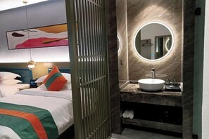 A hotel room with a bed, a bedside table, a nightstand, a wall-mounted mirror, a round mirror, a bathroom vanity with a sink, and a towel rack. - Qianxun Intelligent Hotel Haiyisu (Qibala International Resort) (Menghai)