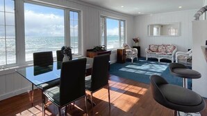 Una habitación amplia con una mesa de vidrio grande y sillas, un rincón de descanso acogedor con un sofá y sillones, y vistas al océano a través de grandes ventanales.
