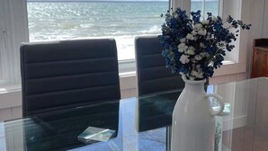 Comedor con mesa y sillas de vidrio, un florero con flores y vista al océano a través de una ventana.