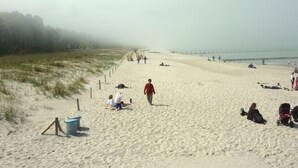 Una platja sorrenca amb gent caminant i asseguda, un moll a la distància i una tanca al costat de la riba.