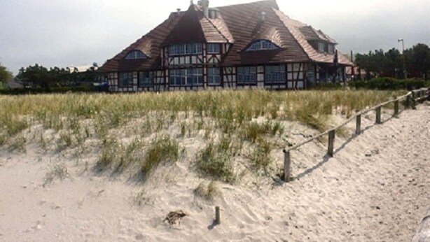 Una casa espaiosa de dos pisos amb un teulat pronunciat, envoltada de dunes de sorra i una tanca de fusta.