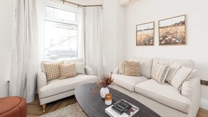 Un salón moderno con un sofá beige, una mesa de centro y un jarrón con flores secas. Hay dos cuadros en la pared y una ventana con cortinas.