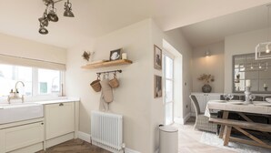 Una cocina moderna con encimera blanca, suelo de madera en espiga y radiador blanco. Hay una zona de comedor con mesa y sillas de madera, y un cubo de basura blanco.