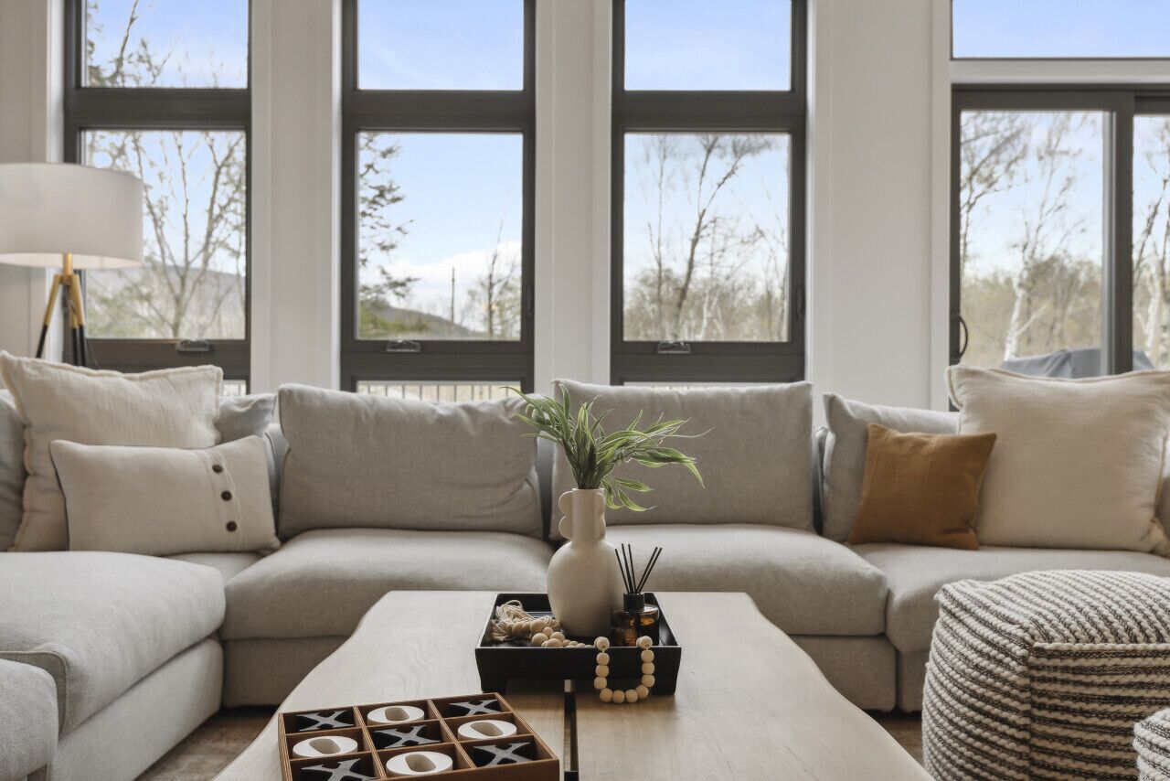 Ein modernes Wohnzimmer mit einem Ecksofa, einem Couchtisch mit Dekoration und großen Fenstern mit Blick auf Bäume.