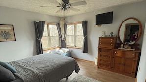 Une chambre à coucher comprenant un lit, une commode, un ventilateur de plafond, une fenêtre avec des rideaux et un téléviseur fixé au mur.