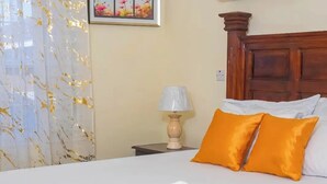 Un llit ben fet amb llençols blancs i dos coixins taronges, un reposacapses de fusta, una taula de nit amb una làmpada i un quadre al mur.