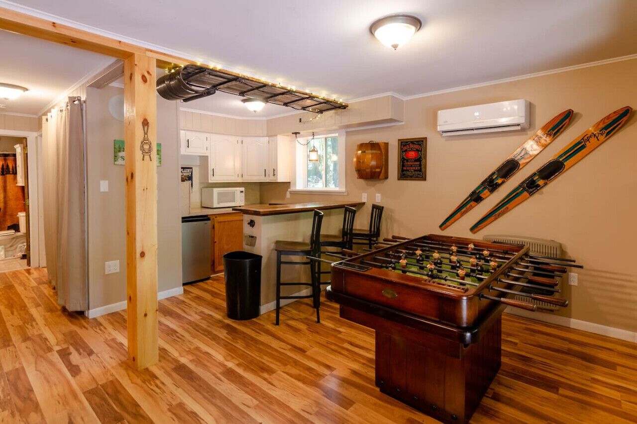 A cozy living room with a foosball table, wooden flooring, a kitchen area with white cabinets, and a mounted ski rack.