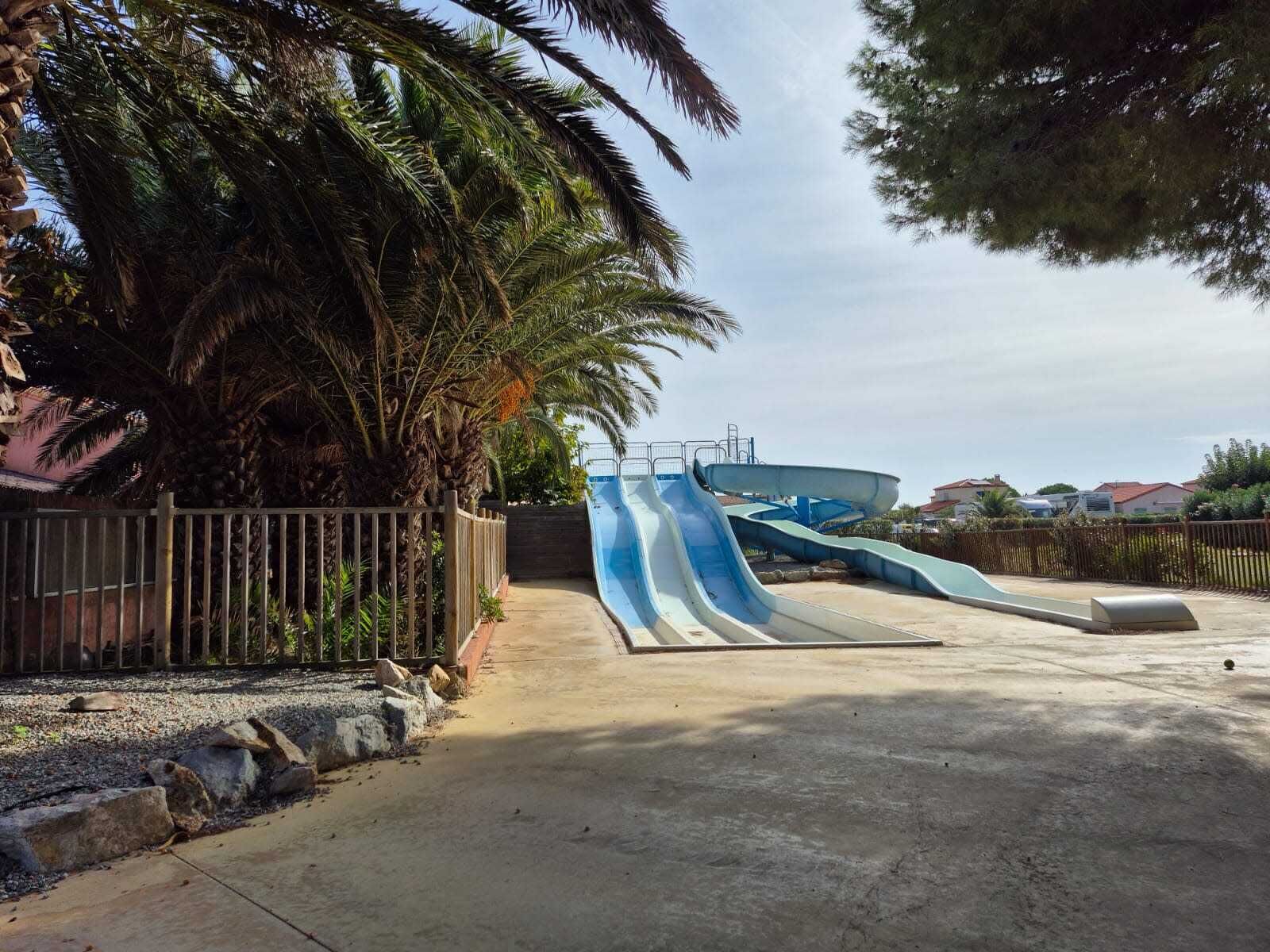 A water park with a blue and white water slide, surrounded by palm trees and a concrete path.