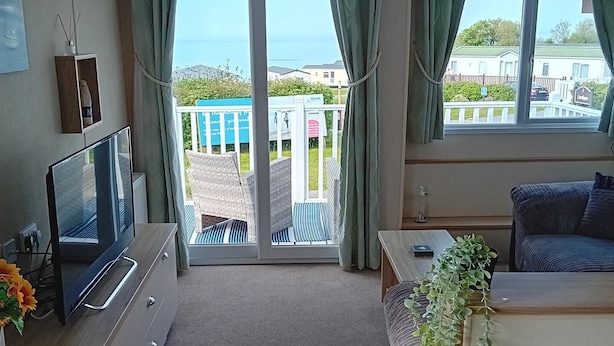 A compact room with a TV, a small desk, a sofa, and a balcony with a view of the sea and buildings. - Seaside Chalet Retreat in New Quay (New Quay)