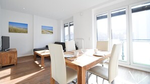 A modern living room with a wooden dining table, white chairs, a sofa, and a TV on a wooden cabinet. - Haus Meeresloft - FW 3 (Norderney)