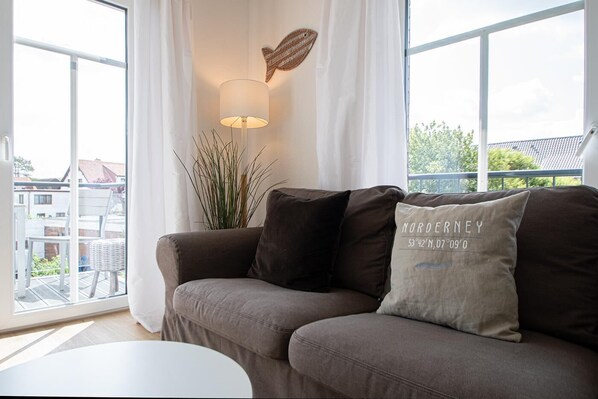 A modern living room with a brown sofa, a white table, and a view of the neighborhood through large windows. - In der Deichresidenz Norderney Finden Sie Sechs Komfortable Luxus-ferienwohnungen, die im Sommer 2012 Fertiggestellt Wurden. Hell und Freundlich Eingerichtet und Hochwertig Ausgestattet Laden die Wohnungen zu Einem Urlaub auf Höchstem Niveau Ein (Norderney)