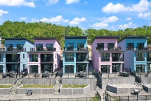 Une rangée de bâtiments modernes aux couleurs vives, dotés de balcons, un mur de soutènement en pierre et, en arrière-plan, une colline boisée.