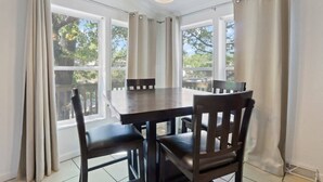 A dining area with a table and chairs, large windows with curtains, and a ceiling light fixture.