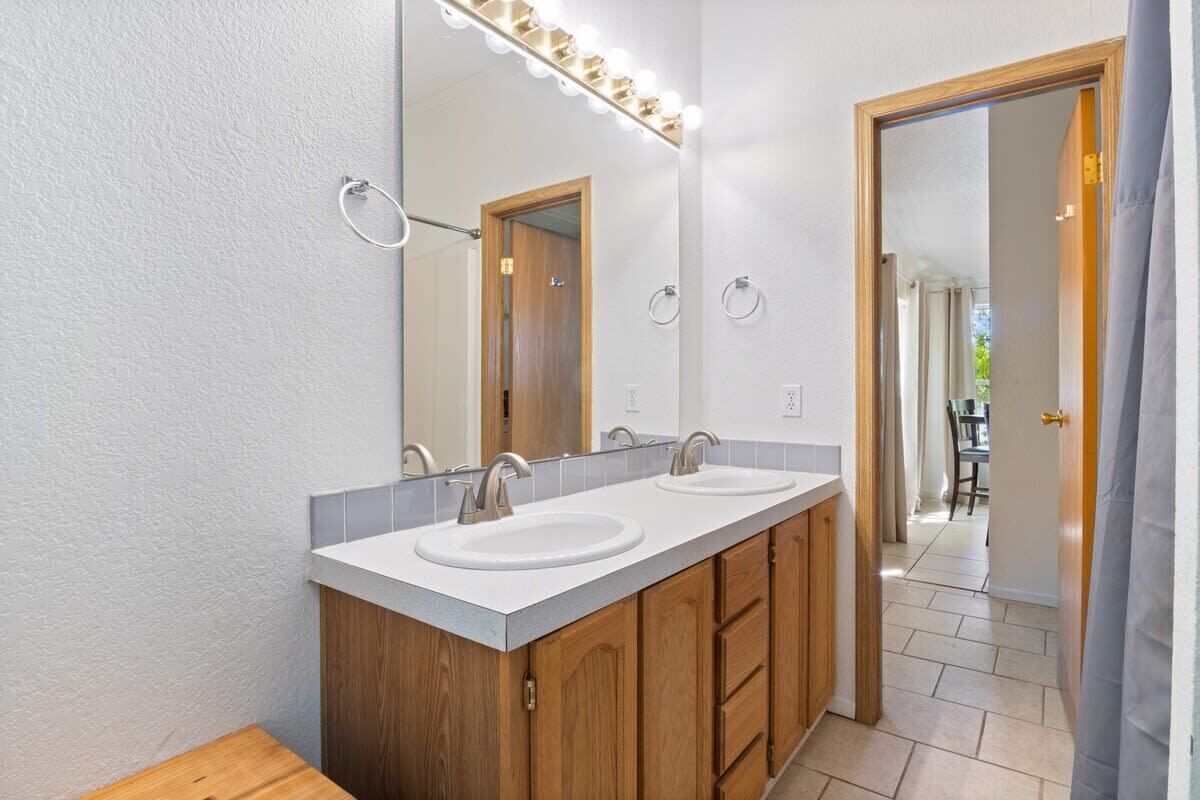 A bathroom with two sinks, wooden cabinets, a large mirror with lights, and a door leading to another room.