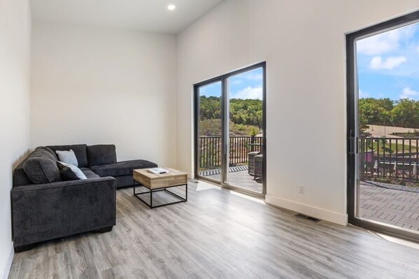 A spacious living room with a grey sofa, a wooden coffee table, and a large sliding glass door leading to an outdoor balcony.