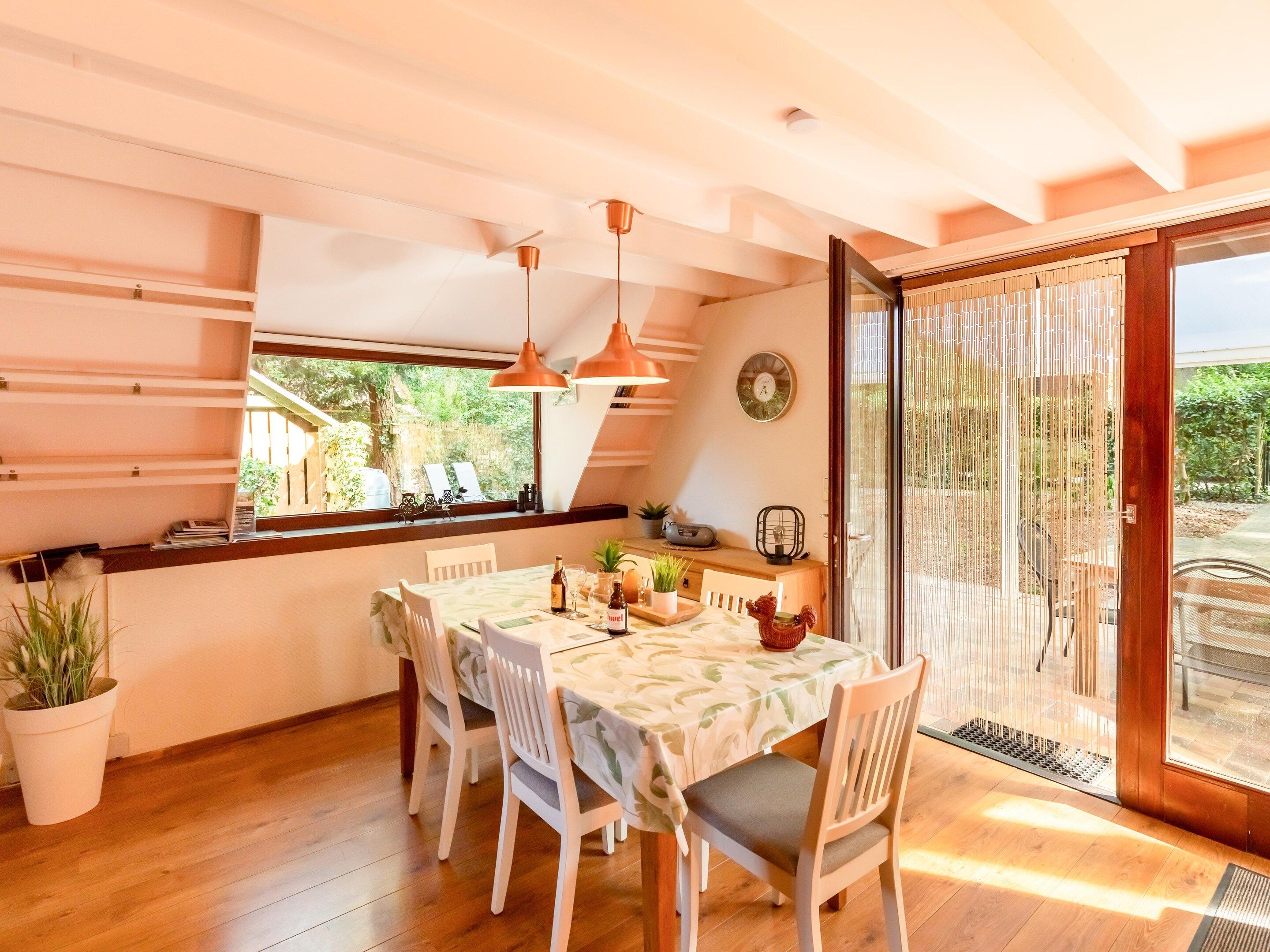 A dining area with a table set for four, wooden chairs, pendant lights, a window with a view of greenery, and a sliding glass door leading outside.