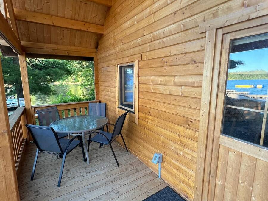 A wooden cabin with a covered patio featuring a glass table and chairs, a view of a lake, and a door with a window.