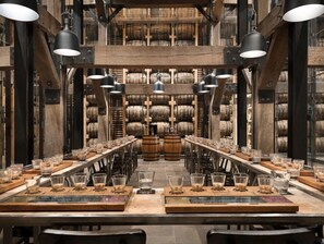 A long table with multiple glasses of whiskey, surrounded by wooden barrels and metal beams.