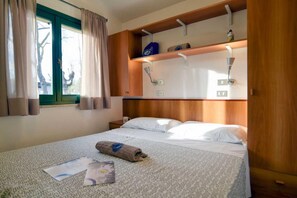 Een net opgemaakt bed met een opgerolde handdoek en een tijdschrift, een raam met gordijnen, en een plank met een lamp en een klein object erop.