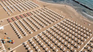 Vista aérea de una playa con hileras de sombrillas y sillas de playa blancas, una orilla arenosa y un rompeolas al fondo.