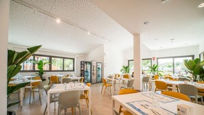 Un comedor amplio con varias mesas y sillas, ventanas grandes y plantas de interior.