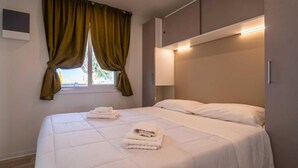 A neatly made bed with white linens and towels, a window with green curtains, and a bedside lamp.