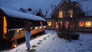 A snow-covered outdoor area with a wooden structure, possibly a sauna, and a house with lit windows in the background. - Holiday Home Bloom 2 (Zingst)