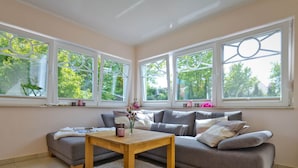 A modern living room with a grey sofa, a wooden coffee table, and a white rug. There are large windows with decorative frames and a view of greenery outside. - Holiday Home Bloom 2 (Zingst)