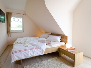 A bedroom with a sloped ceiling, a bed with white bedding, a nightstand with a lamp, and a window with curtains. - Holiday Home Bloom 2 (Zingst)