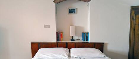 A neatly made bed with two pillows and two folded towels, a bedside lamp, and a small shelf with books and a framed picture.