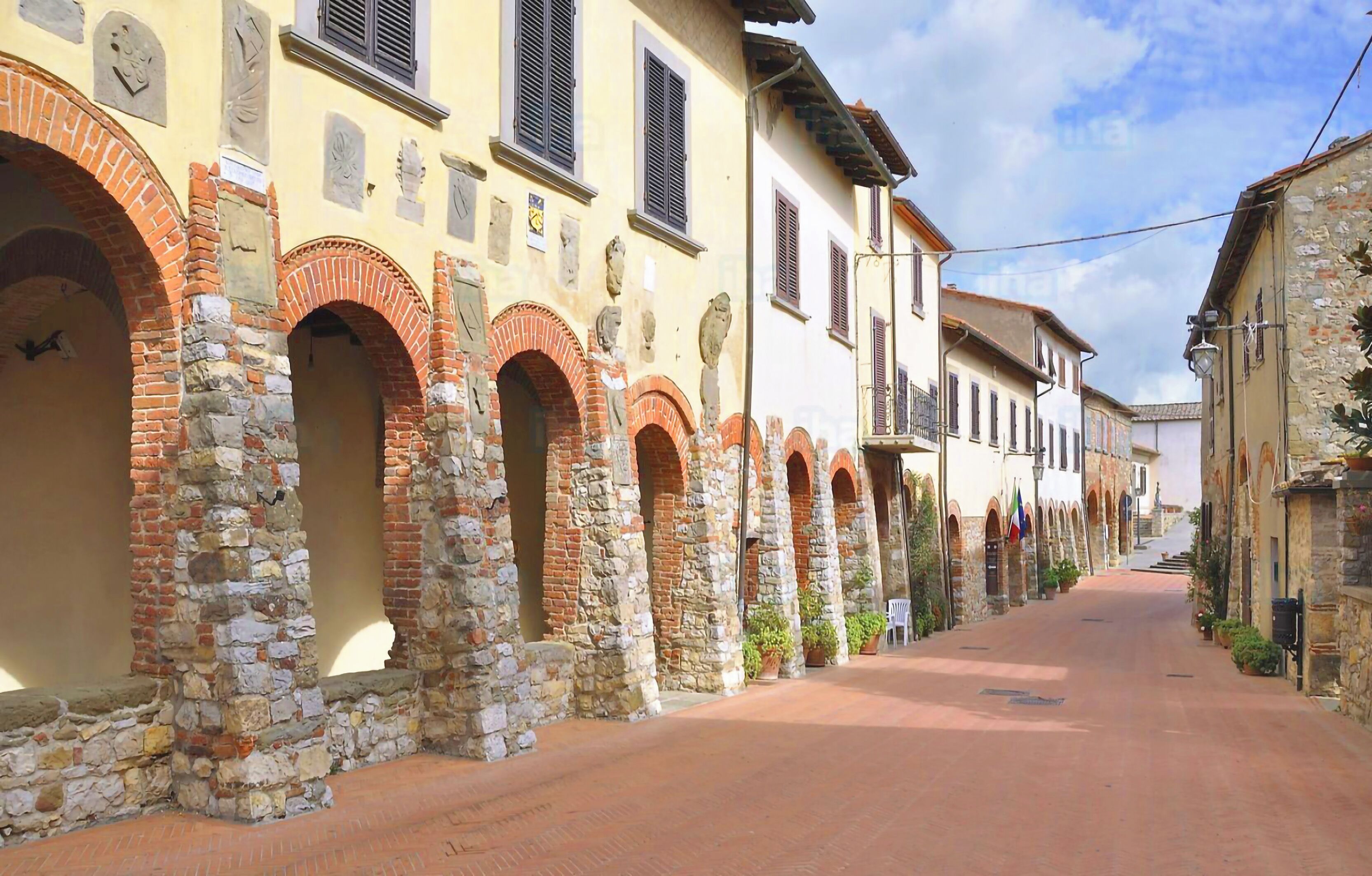 Una calle con edificios históricos que presentan fachadas de ladrillo y piedra con arcos, un camino pavimentado y plantas en macetas a lo largo de la acera.