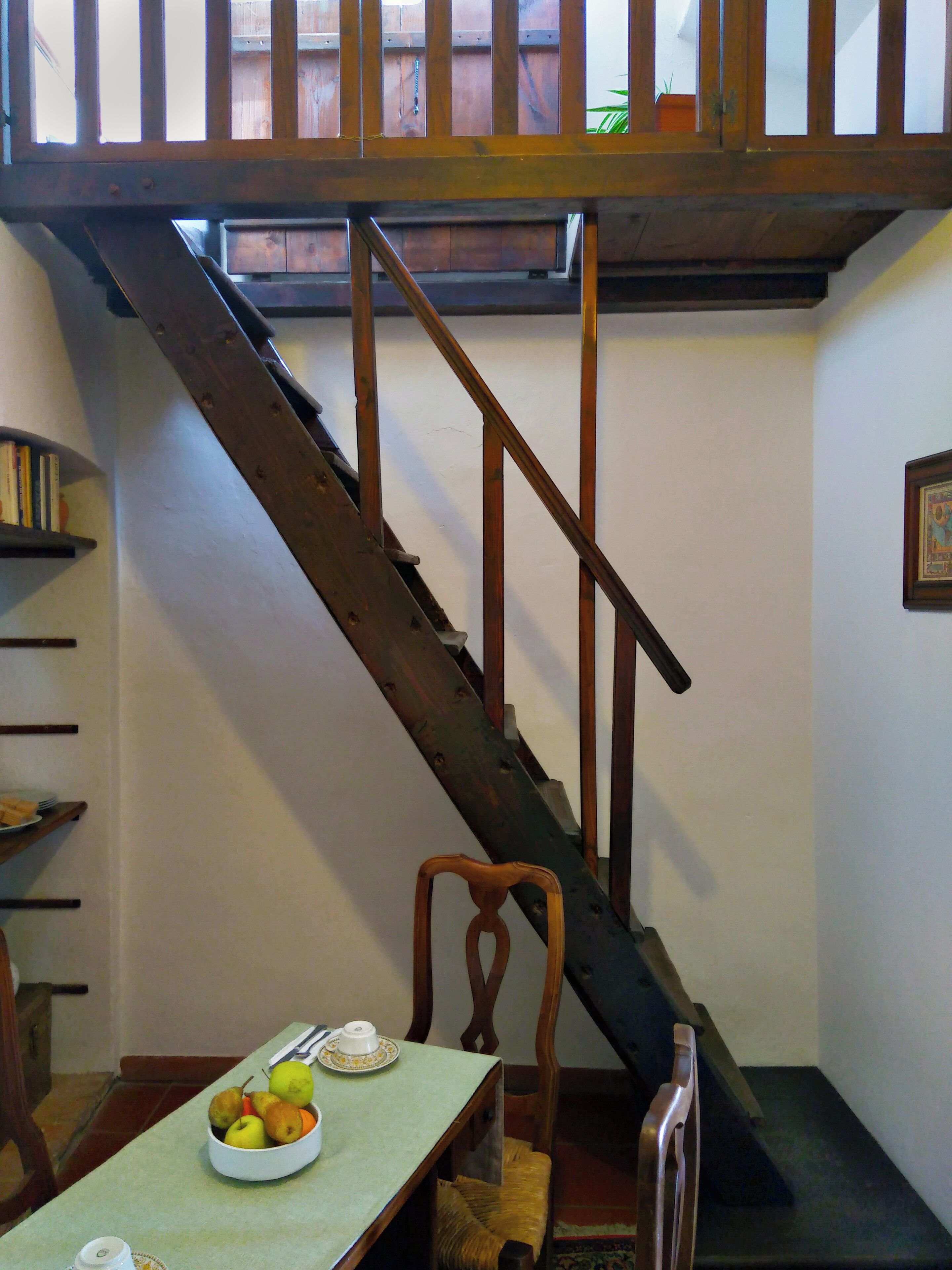 A staircase with wooden railings leading to an upper level. A table with a bowl of fruit and a teapot is positioned at the bottom of the stairs.