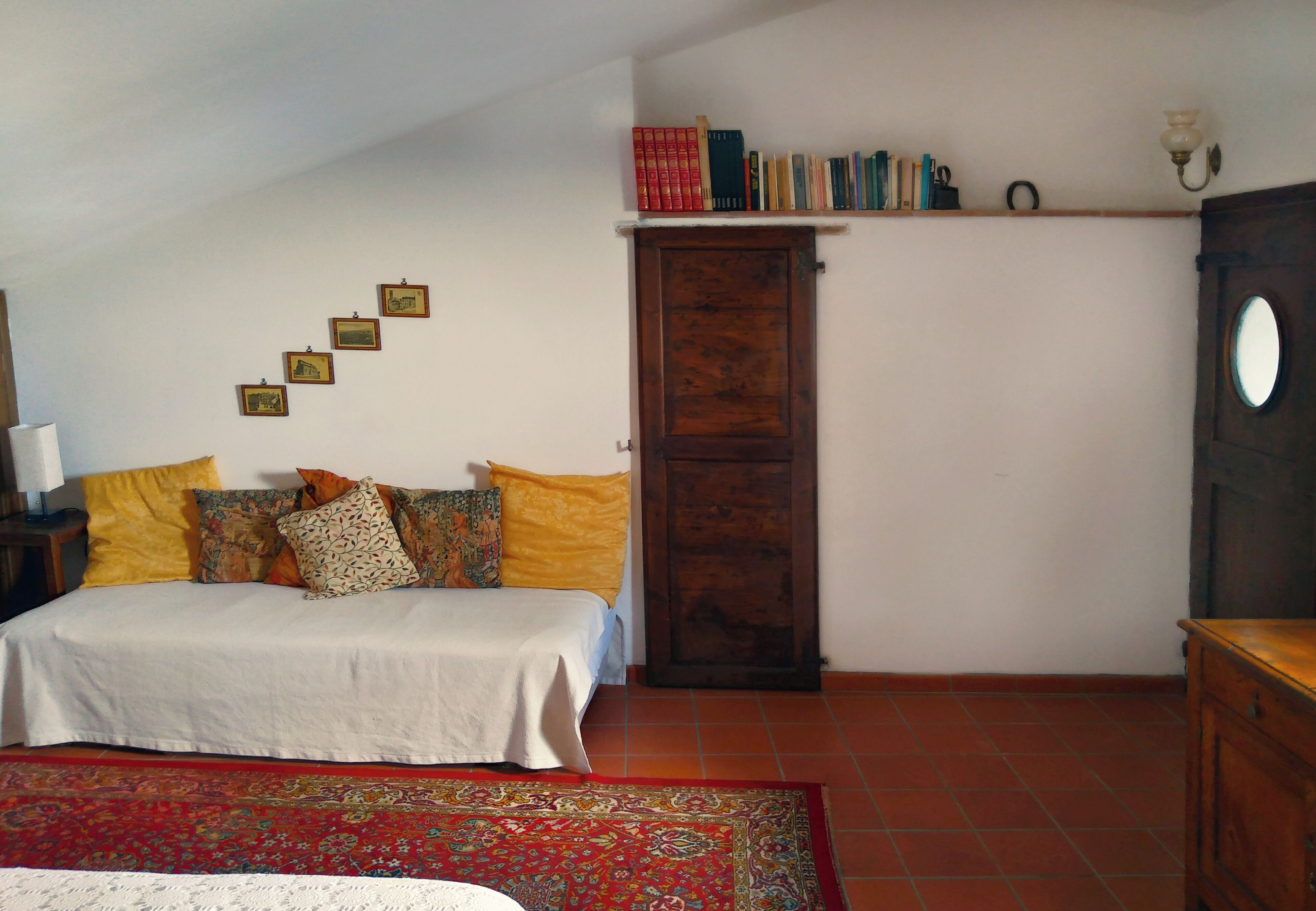 A bedroom with a bed, a wooden wardrobe, a shelf with books, and a patterned rug.