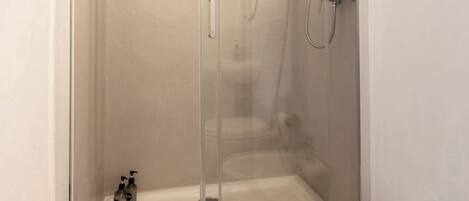 Ducha con mampara de vidrio, cabezal de ducha de mano, lavamanos blanco y una toalla en el suelo.