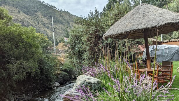 A riverside setting with a thatched-roof gazebo, a dining area with wooden furniture, and a mountainous backdrop. - Full Moon Eco Lodge (Ollantaytambo)