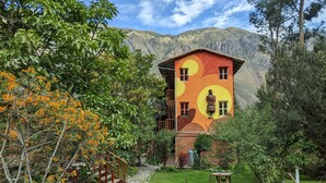 A house with a colorful exterior, surrounded by a lush garden and a mountain backdrop. - Full Moon Eco Lodge (Ollantaytambo)