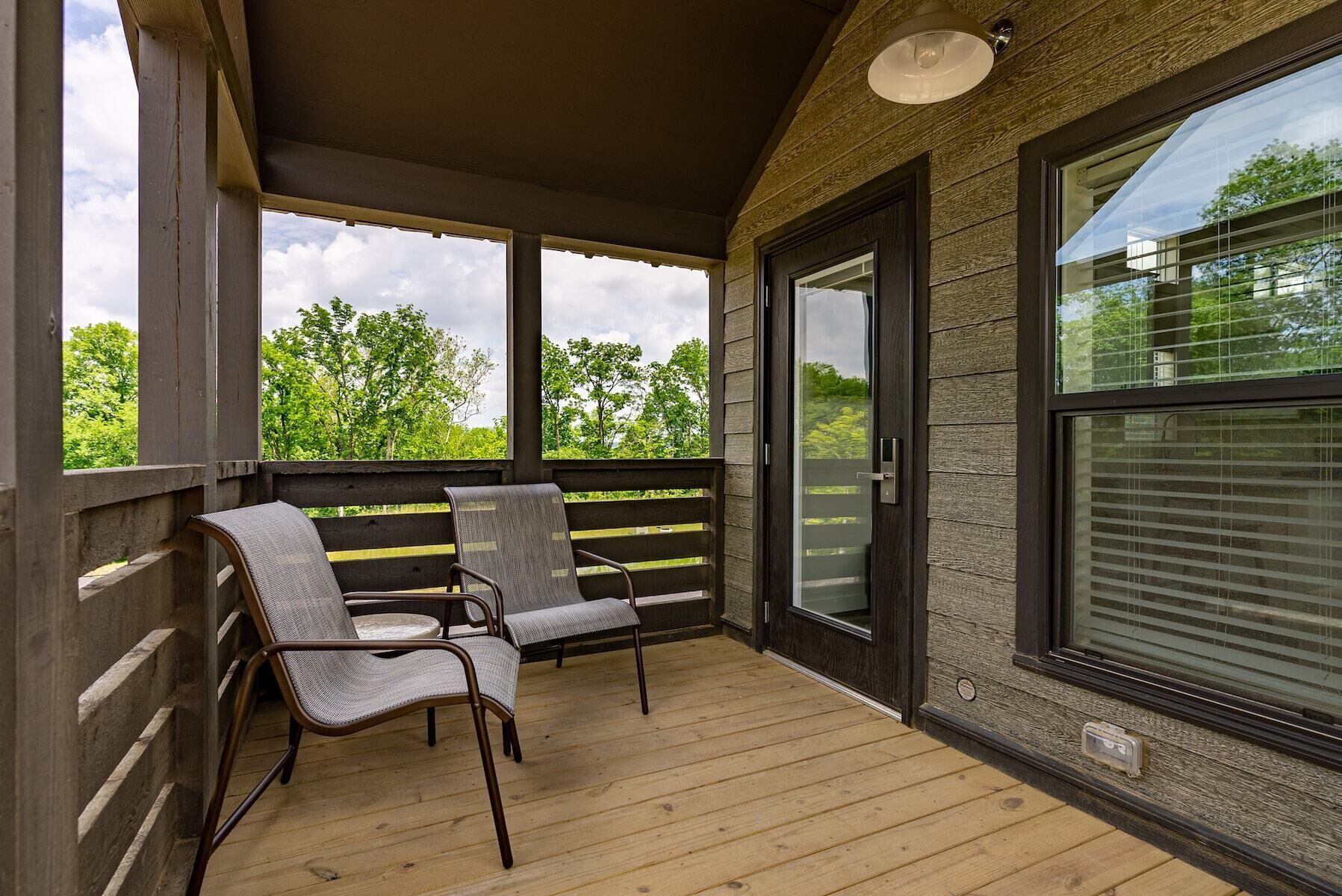 A covered outdoor area with two chairs and a wooden floor, large windows, and a view of trees.