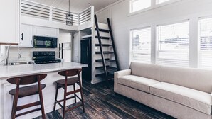 A modern kitchen with white cabinets, a black refrigerator, a microwave, and a stainless steel sink. There is a wooden ladder leaning against the wall, a beige sofa, and a window with blinds. - Cincinnati Camp Cedar (Mason)