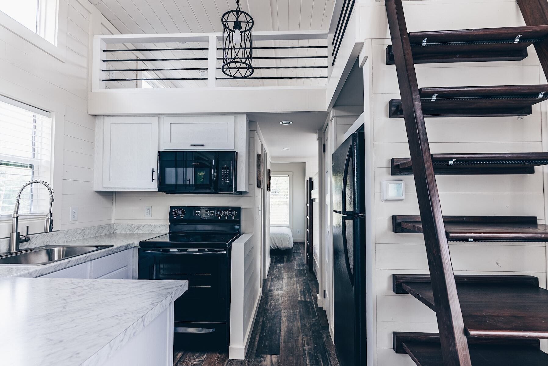 Una cocina moderna con armarios blancos, un microondas negro, una estufa y un refrigerador. A la derecha se ve una escalera de madera y a la izquierda, una ventana.