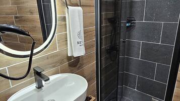 Un baño moderno con piso de madera, un lavamanos blanco con gabinete de madera, cerramiento de ducha de vidrio y un espejo con luz.