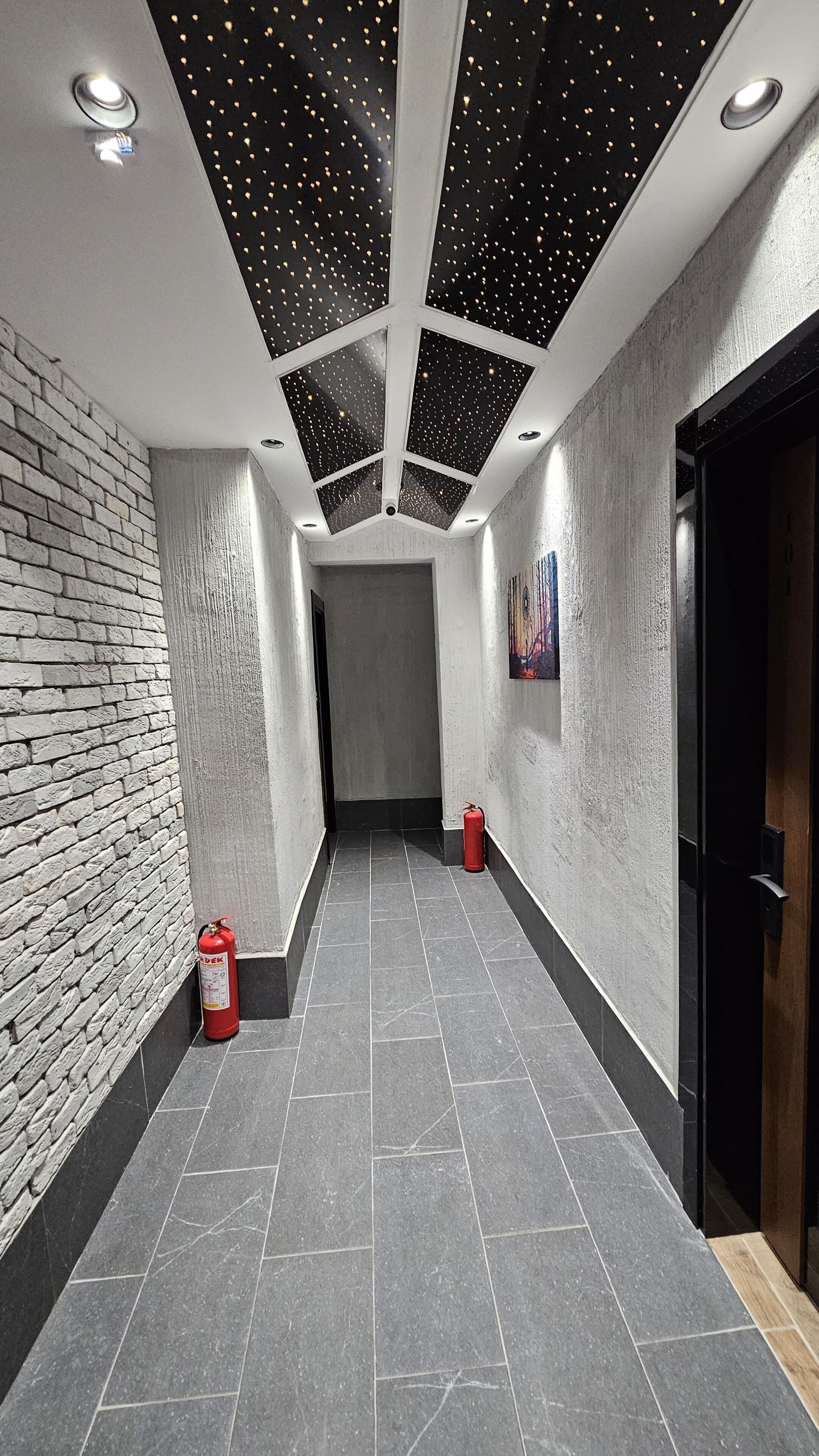 A hallway with a brick wall on one side, a tiled floor, and a ceiling with star-like lights. There is a red fire extinguisher on the floor.