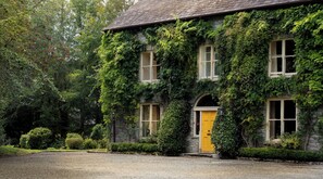 A stone building with a yellow door and windows, covered in green ivy, surrounded by trees and a gravel driveway.