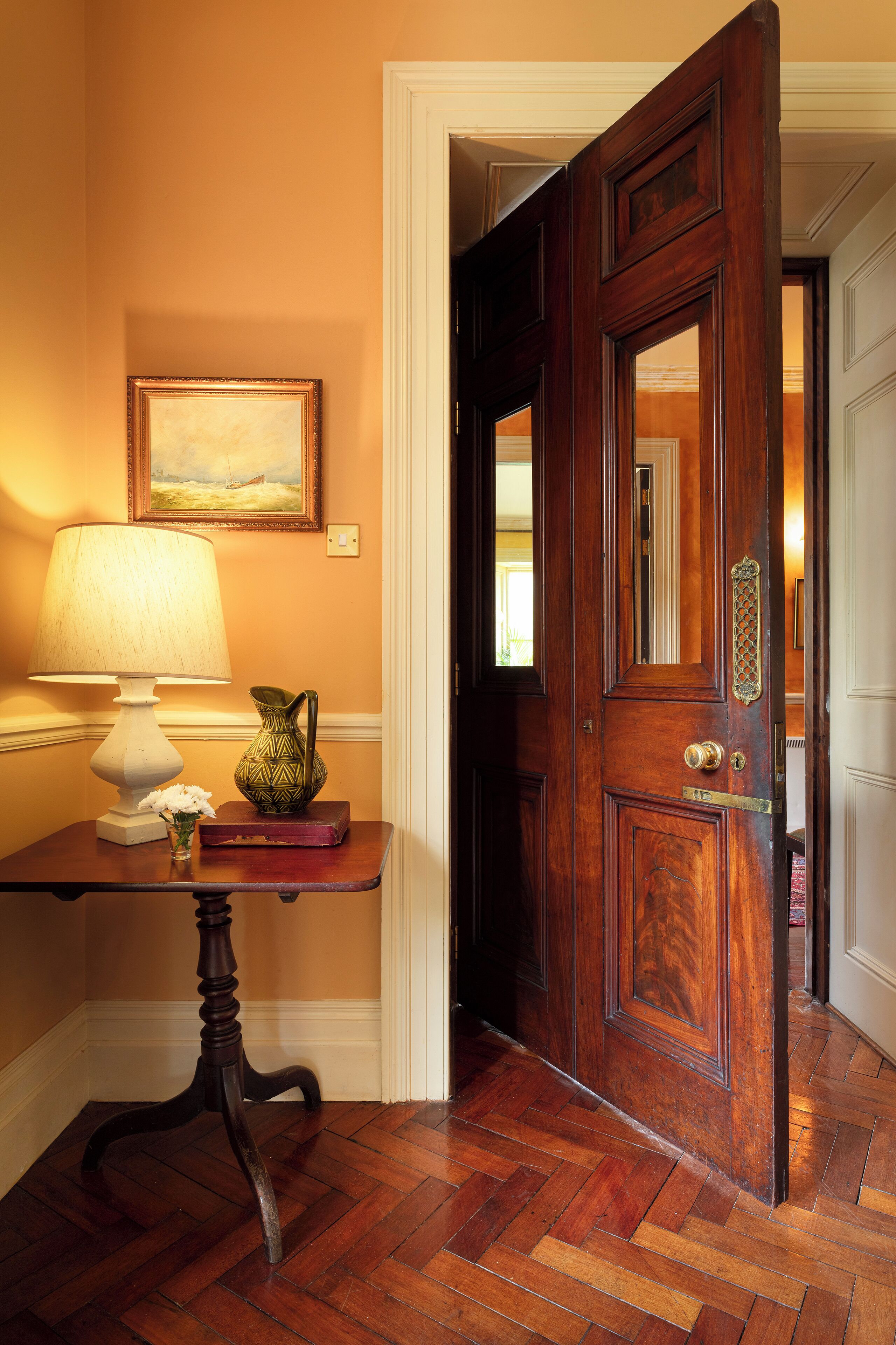 Una puerta de madera con manija de latón, un cuadro enmarcado en la pared y una mesa con una lámpara y un jarrón.