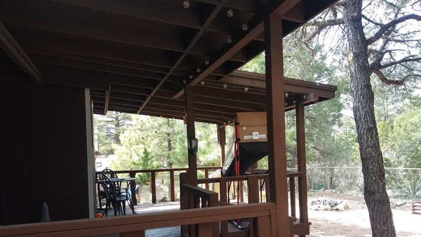 Eine Holzterrasse mit überdachter Sitzgelegenheit, einem Grill und Blick auf Bäume und einen Zaun.