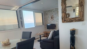 Ein modernes Wohnzimmer mit Sofa, Couchtisch und Fernseher. Ein großes Fenster bietet Blick auf den Ozean.