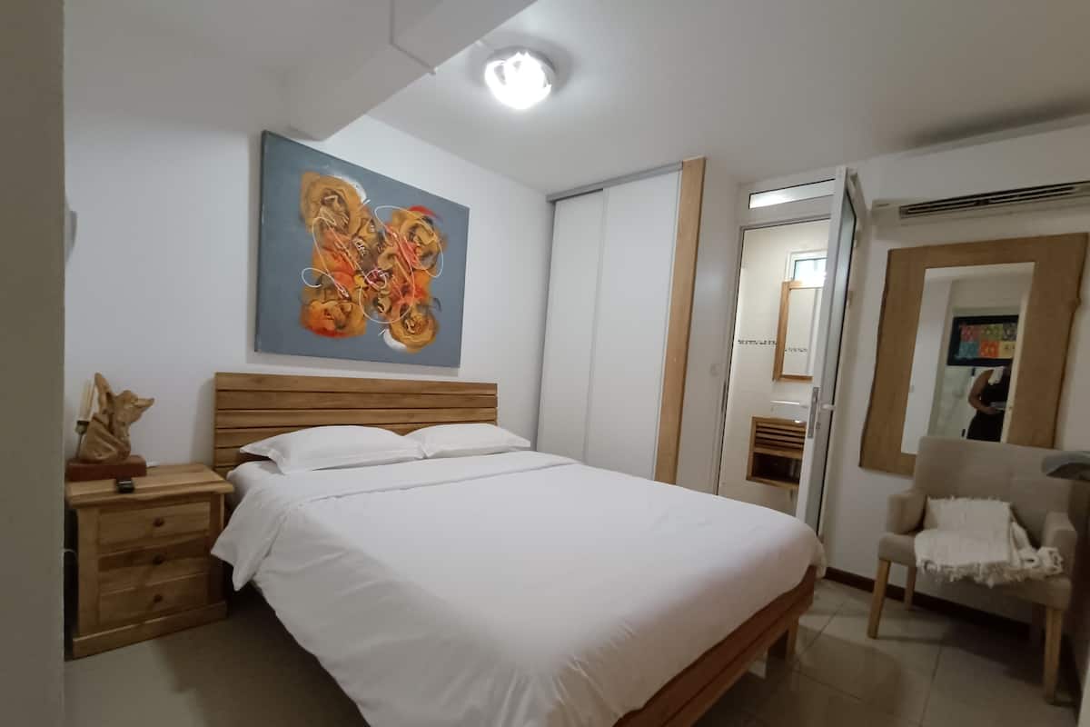 A bedroom with a wooden bed, white bedding, a nightstand with a lamp, a painting on the wall, and a chair with a cushion.