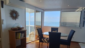 Ein Zimmer mit Glastisch und Stühlen, einem Holzregal und Meerblick durch große Fenster.